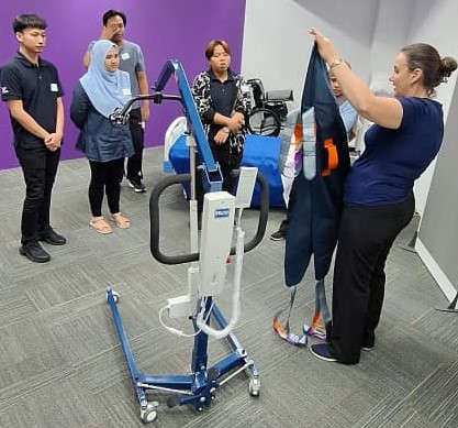 Woman demonstrates a blue patient lift with a sling while four people observe in a clinical room.