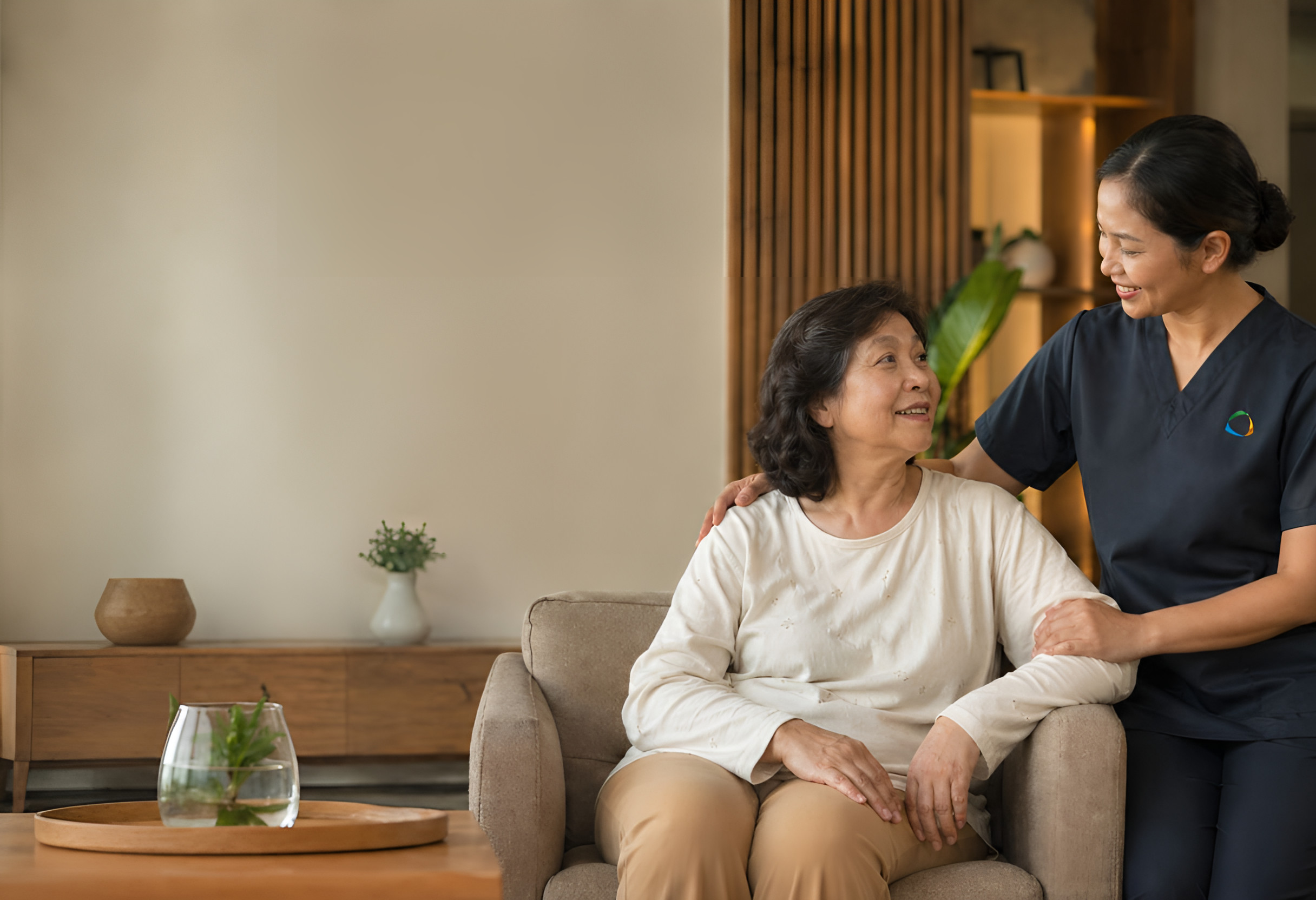 Caregiver in navy scrubs smiles beside an older woman seated in a living-room armchair.