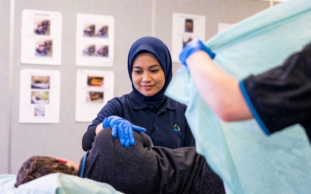 Healthcare workers in blue gloves assist a person on a bed while holding a turquoise drape.